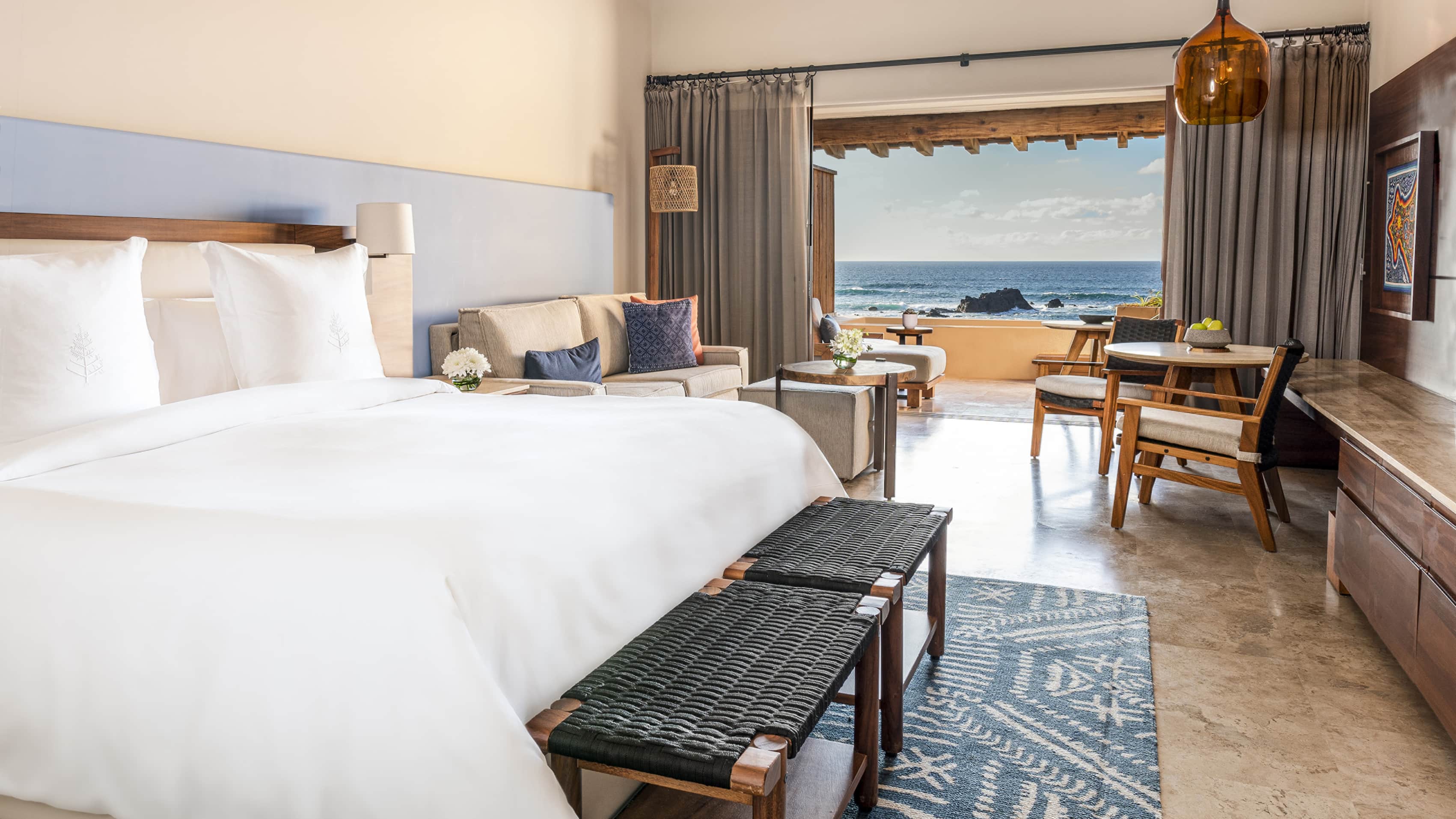 Luxurious ocean-view bedroom at Four Seasons Resort Punta Mita, a top MICE venue in Mexico for meetings, incentives, conventions, and exhibitions, featuring a king-size bed with white linens, blue-patterned rug, wooden benches, cozy seating area with sofa and chairs, fresh flowers, and balcony overlooking the Pacific Ocean and rocky coastline ideal for relaxing during incentive travel or corporate events in Riviera Nayarit.